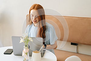 Medium shot of positive attractive young woman student working on project using laptop sitting at desk in cozy cafe