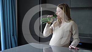 Medium shot pregnant young woman drinking beer from bottle in kitchen at home. Portrait of Caucasian expectant looking