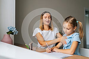 Medium shot of pretty primary daughter doing homework with young mother sitting at home table by window.