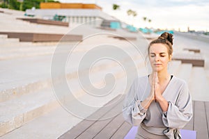 Medium shot of relaxed Caucasian young woman practicing yoga performing namaste pose with closed eyes outside in city