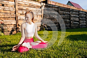 Medium shot of serene pretty young woman sitting on green grass with closed eyes in lotus position holding hands in knee