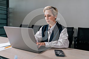 Medium shot of serious professional mature business woman using typing on laptop computer sitting at workplace desk.