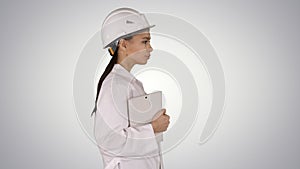 Attractive Hispanic woman in white lab coat and white safety hard hat walking holding notebook or tablet on gradient