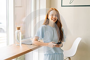 Medium shot of smiling charming young woman holding mobile phone, typing online messages standing by window in cozy
