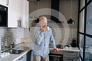 Medium shot of thoughtful gray-haired mature adult male drinking hot coffee and looking through window in morning at