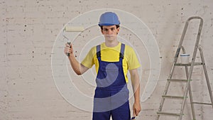 Medium shot of a young construction worker standing in the room under renovation, choosing between a rolloer and a brush