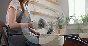 Medium shot of young female potter in pottery workshop. Woman in apron is drying ceramic blank on rotating wheel. Slow