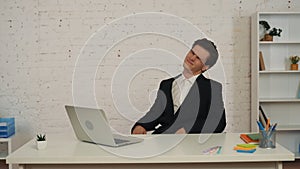 Medium shot of a young man sitting at the table, working on a laptop, stretching his neck that is hurting after a long