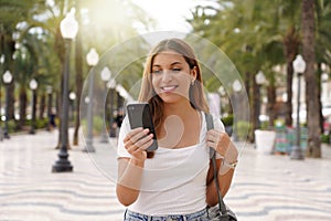 Medium shot young smiling woman uses smartphone reads news from social networks searches text sends mails online strolls at street