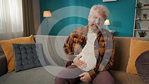 Medium-sized shot of an eldery, retired man, senior citizen sitting on a couch, sofa, feeling severe abdominal pain