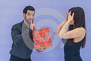 Medium studio shot on blue background of a cuban bearded handsome man holding a colorful present, puting a finger on his