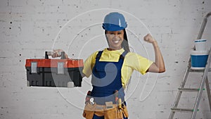 Medium video of a dark-skinned young female construction worker standing in the room, wearing a tool belt, raising a