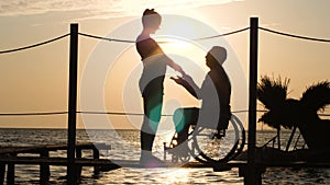 Meeting of slender girl with disabled guy on wheelchair at bright sunset standing on pier near water
