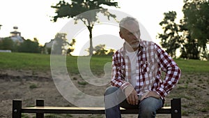 Melancholic elderly male sitting on bench in park and thinking about problems