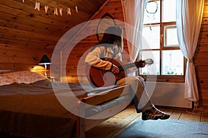 Melancholic lonely woman sits on bed with guitar in hands and looks out window in cozy wooden house