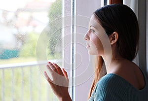 Melancholic sad young woman looking through the window at home in a rainy day, focus on model eyes, indoor photo