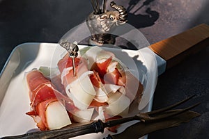 melon in jamon. gourmet cuisine: melon with ham. Summer snack with sweet melon and ham on a dark background. hard light