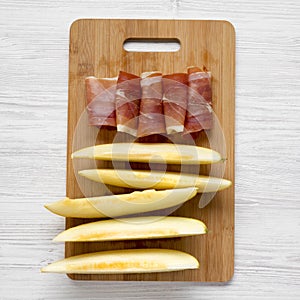 Melon slices and prosciutto on bamboo board over white wooden background, overhead view.