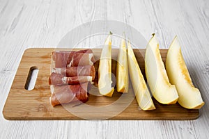 Melon slices with prosciutto on bamboo board over white wooden background, side view.