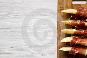 Melon slices wrapped in prosciutto on bamboo board over white wooden background, top view. Close-up. From above, overhead, flat la