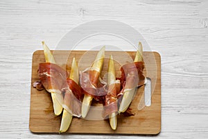 Melon slices wrapped in prosciutto on bamboo board over white wooden table, overhead view. From above, top view.