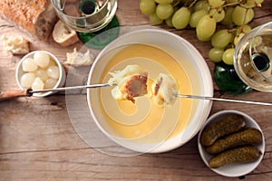 Melted cheese fondue with bread on long forks, pickles, grapes and white wine on a rustic wooden table, copy space, high angle