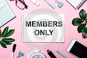 MEMBERS ONLY is written in a white notebook on a pink background surrounded by business accessories and green leaves photo