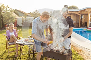 Men grilling meat while women are  sitting and relaxing by the pool at backyard barbecue party