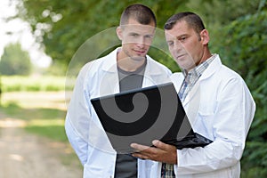 Men oudoors wearing labcoats looking at laptop