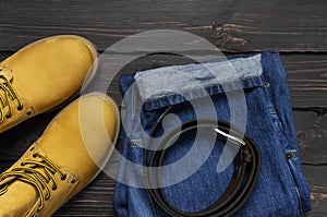 Men`s casual wear, yellow work boots from natural nubuck leather, blue jeans and brown belt on dark wooden background top view