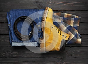 Men`s casual wear, yellow work boots from natural nubuck leather, blue jeans, checkered shirt and brown belt on dark wooden