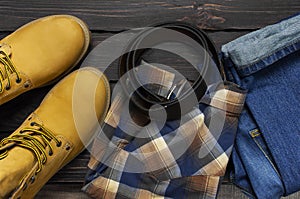 Men`s casual wear, yellow work boots from natural nubuck leather, blue jeans, checkered shirt and brown belt on dark wooden