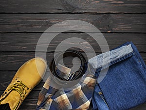 Men`s casual wear, yellow work boots from natural nubuck leather, blue jeans, checkered shirt and brown belt on dark wooden