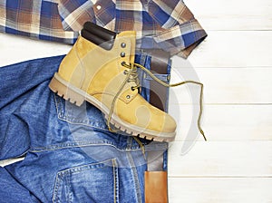 Men`s casual wear, yellow work boots from natural nubuck leather, blue jeans, checkered shirt and brown belt on wooden white