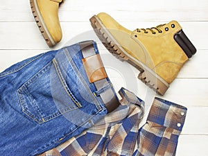 Men`s casual wear, yellow work boots from natural nubuck leather, blue jeans, checkered shirt and brown belt on wooden white