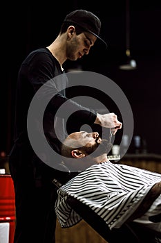 Men`s fashion. The barber dressed in a black clothes scissors beard of brutal man in the stylish barbershop