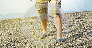 Men& x27;s feet in flip-flops and shorts, a man standing on the beach on a pebble beach, sunrise in the morning