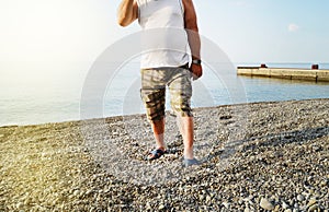 Men& x27;s feet in flip-flops and shorts, a man standing on the beach on a pebble beach, sunrise in the morning