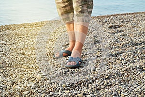 Men& x27;s feet in flip-flops and shorts, a man standing on the beach on a pebble beach, sunrise in the morning