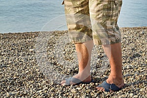 Men& x27;s feet in flip-flops and shorts, a man standing on the beach on a pebble beach, sunrise in the morning