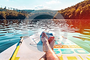 Men`s feet on a paddleboard. First person view. Close-up of legs on surf. Relax and travel on the lake at hot summer day