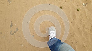 men's feet in sneakers, a man in jeans walking on the sand, top view from the first person.