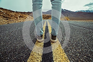 Men`s feet standing on asphalt desert road with yellow marking lines. Man wearing sneakers and jeans.