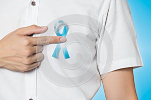 Men`s healthcare concept - close up of male hand pointing to light blue ribbon for prostate cancer at left chest on blue