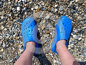 Men`s legs, feet in blue rubber slippers against the background of pebbles and sand on a vacation on the beach in a warm tropical