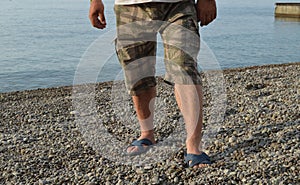 Men`s legs in flip-flops and shorts, a man standing on the beach on a pebble beach