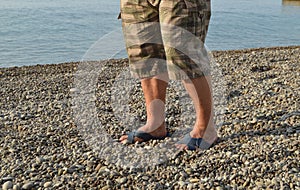 Men`s legs in flip-flops and shorts, a man standing on the beach on a pebble beach