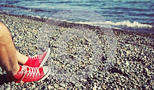 Men`s legs in shorts and red sneakers on a pebbly beach by the sea on a sunny day.