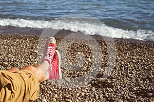 Men`s legs in shorts and red sneakers on a pebbly beach by the sea on a sunny day.