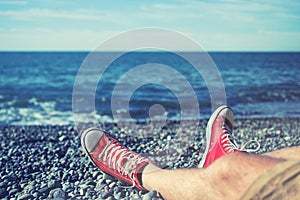 Men`s legs in shorts and red sneakers on a pebbly beach by the sea on a sunny day.